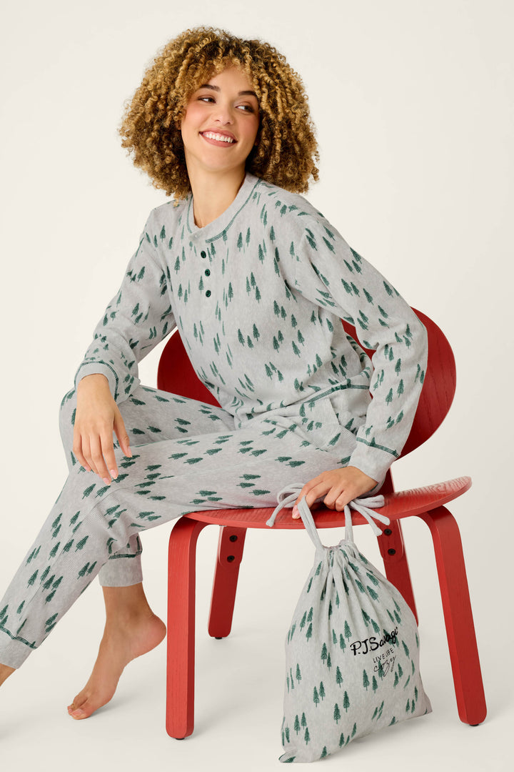 A person wearing the P.J. Salvage Trees Print 3-Piece PJ Gift Set sits on a red chair, smiling and holding the coordinating drawstring bag. The background is plain and light-colored.