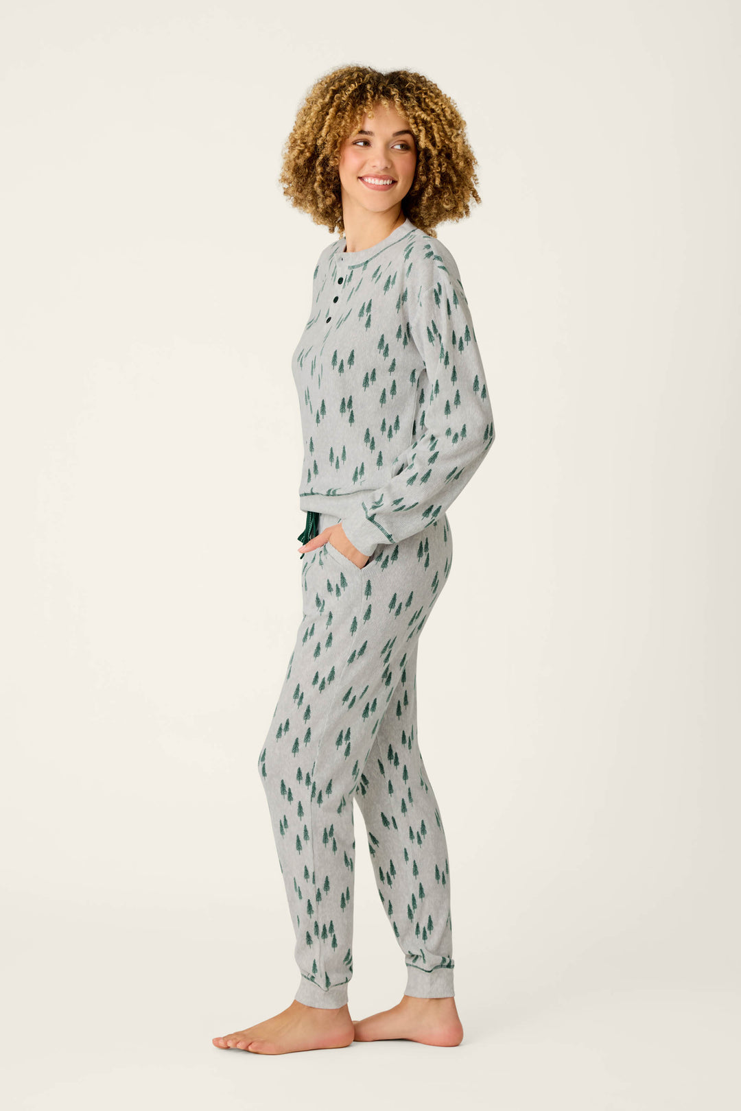 A woman stands barefoot in profile, smiling and looking to the side, wearing the P.J. Salvage Trees Print 3-Piece PJ Gift Set—an elegant light gray pajama set adorned with a green tree pattern—against a plain light background.