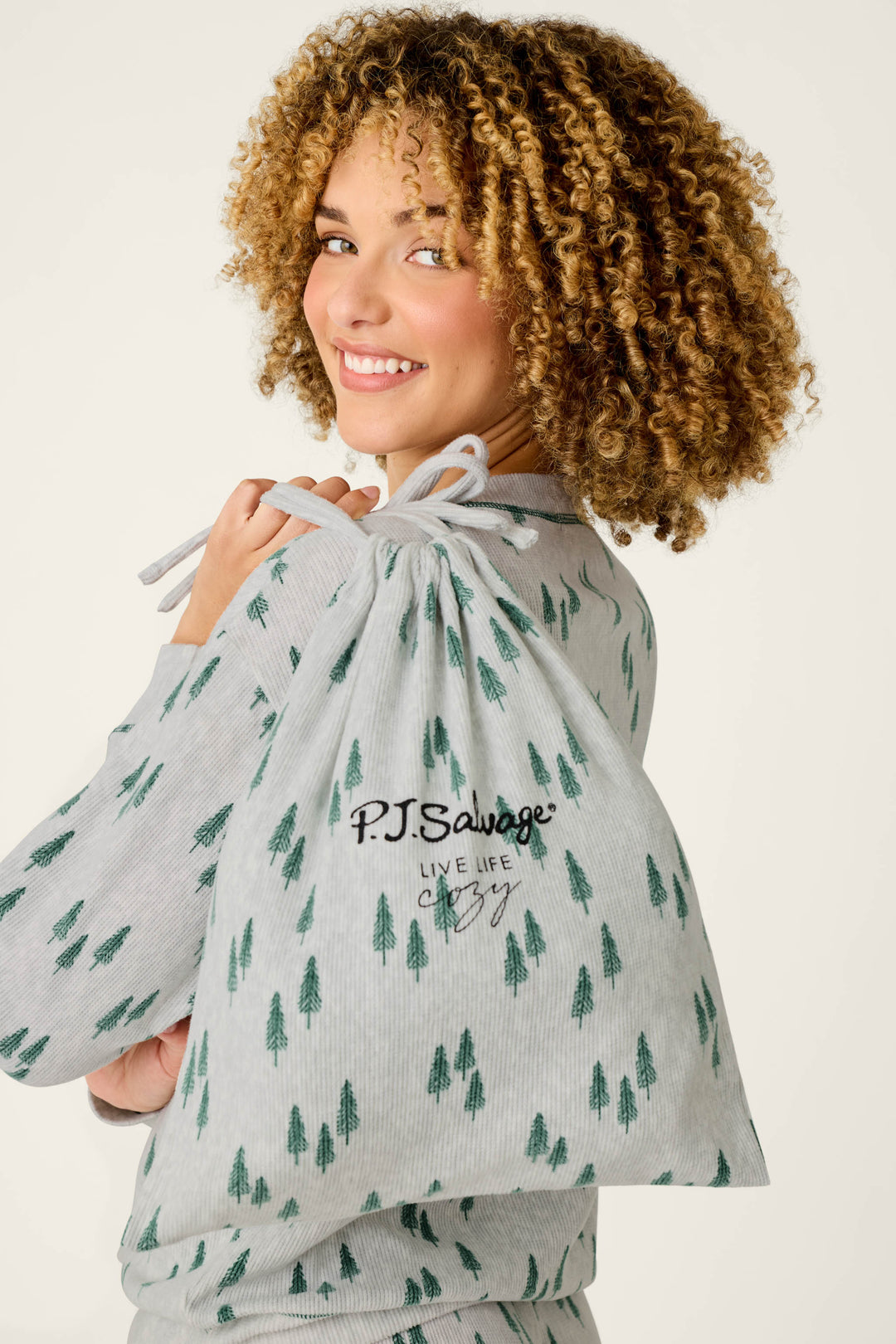 A smiling person with curly hair wears P.J. Salvage’s Trees Print 3-Piece PJ Gift Set, featuring light pajamas patterned with green trees, and carries a matching drawstring bag over their shoulder that reads “LIVE LIFE cozy.”.