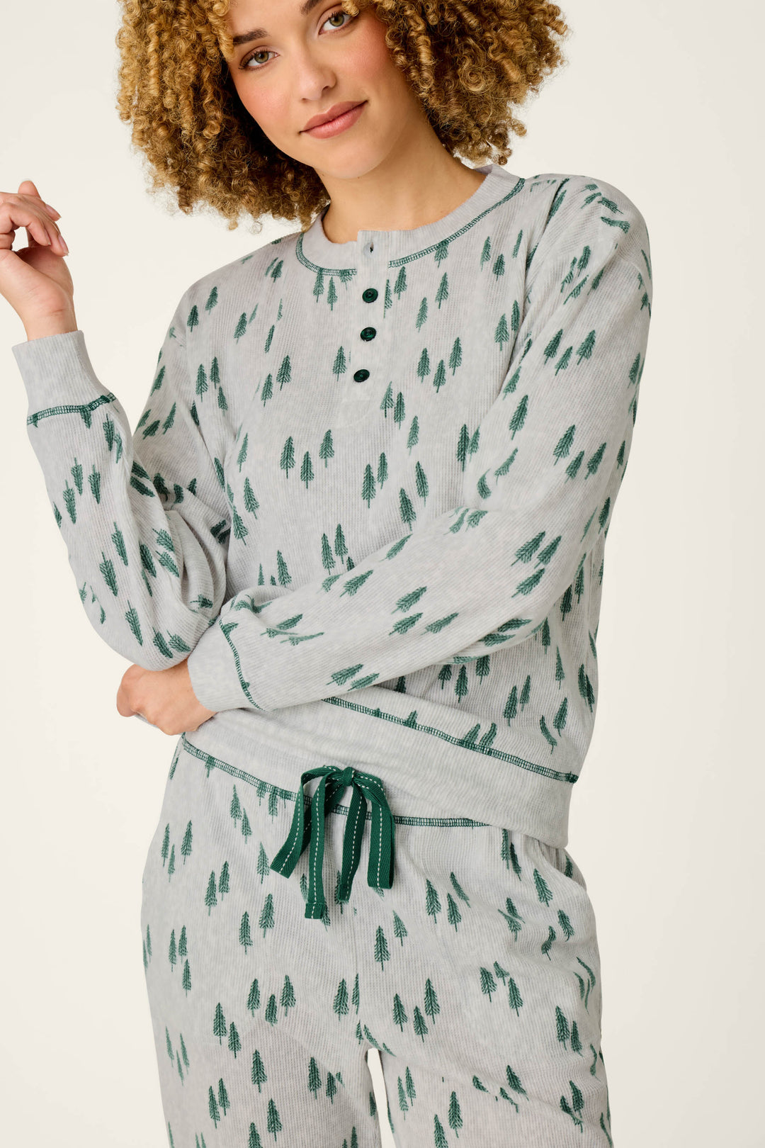A person with curly hair is smiling while wearing the P.J. Salvage Trees Print 3-Piece PJ Gift Set, which features a matching light gray pajama set adorned with a green pine tree pattern. The top includes buttons at the collar, and the pants are finished with a green drawstring.