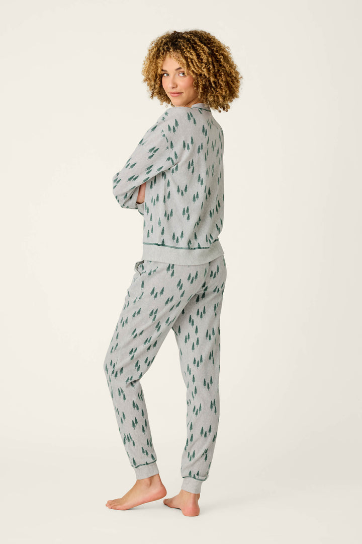 A person with curly hair stands barefoot, turned slightly to the side, wearing the P.J. Salvage Trees Print 3-Piece PJ Gift Set—matching gray pajamas featuring a green tree pattern—against a plain light background.
