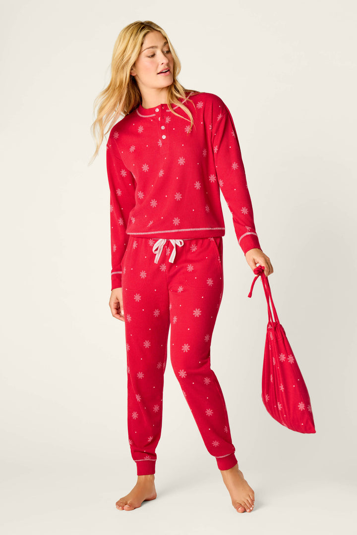 A person stands barefoot against a plain light background, wearing the P.J. Salvage Snowflakes Print 3-Piece PJ Gift Set and holding a matching red bag.