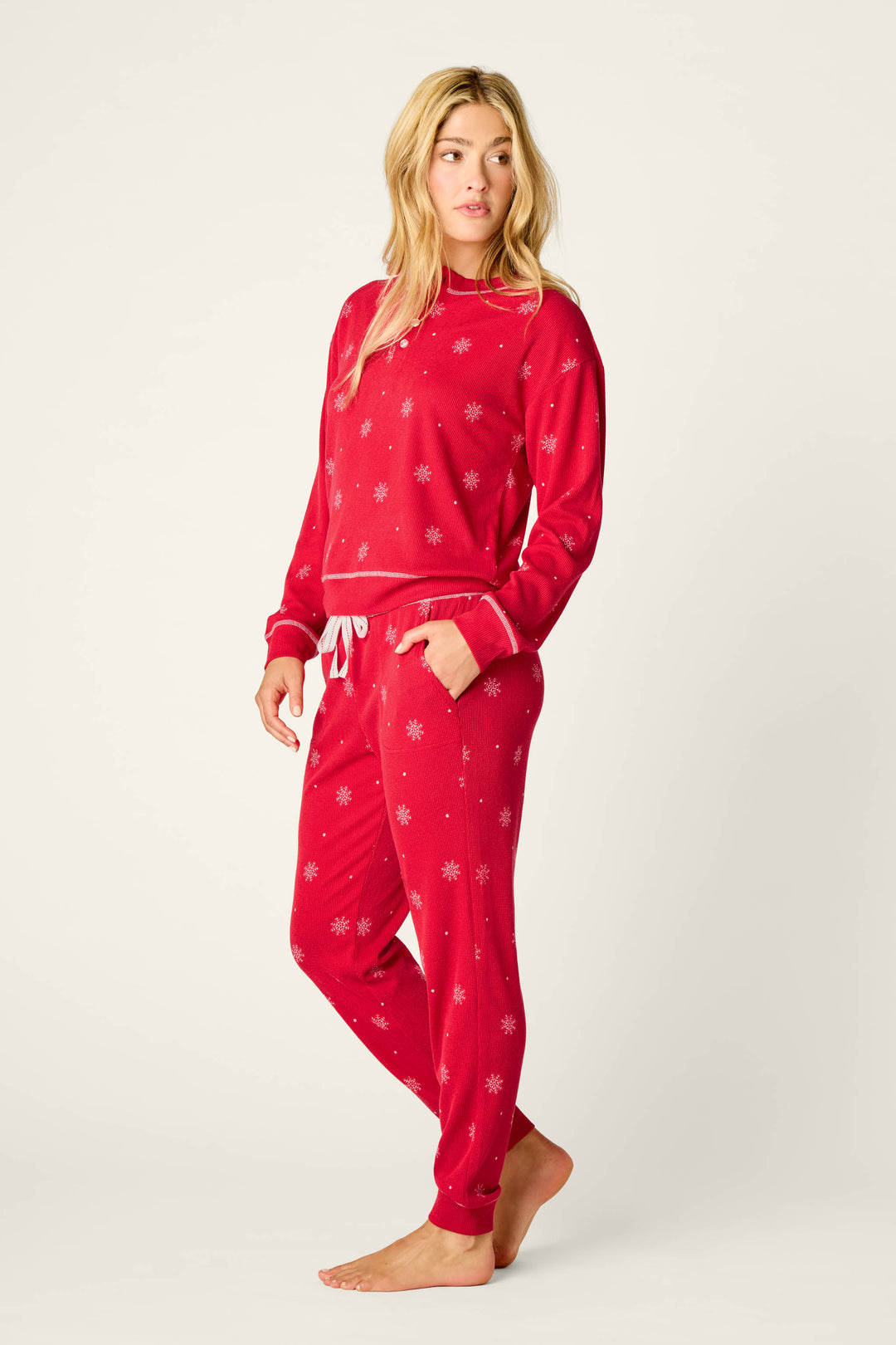A woman stands barefoot, wearing the P.J. Salvage Snowflakes Print 3-Piece PJ Gift Set featuring a matching red design with white snowflakes. She has long blonde hair and a relaxed expression, with one hand in her pocket and the other resting by her side.