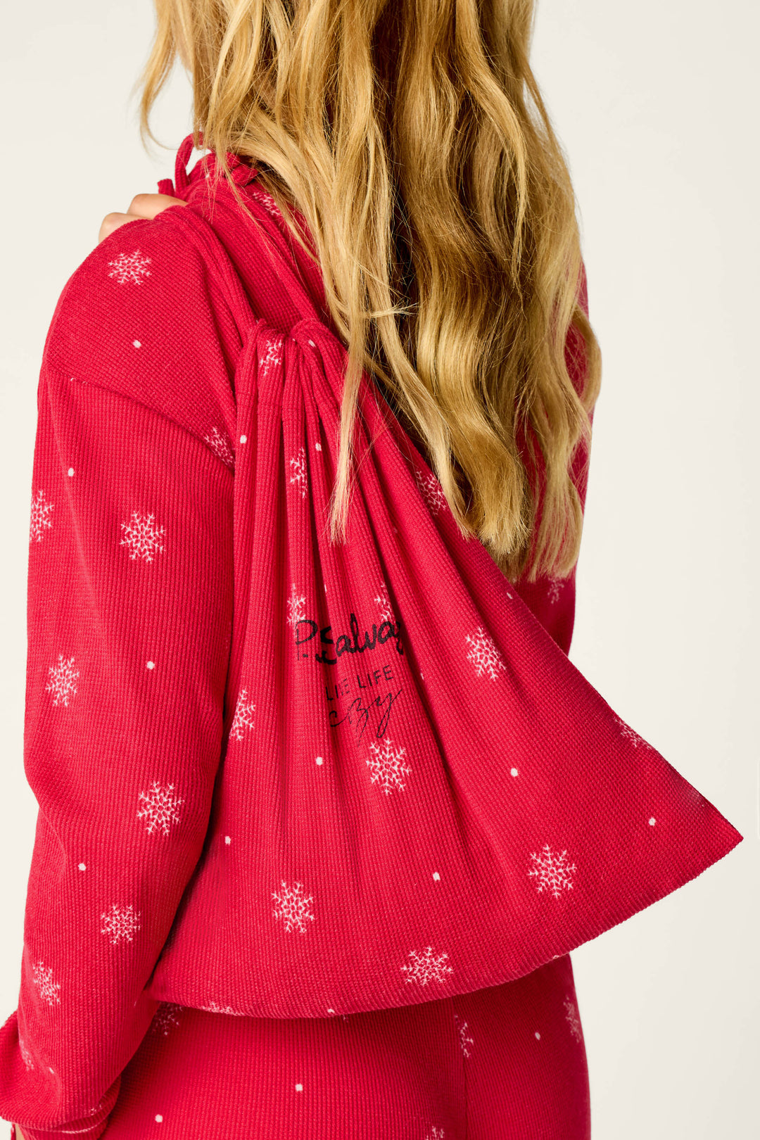 A person with long blonde hair wears P.J. Salvage’s Snowflakes Print 3-Piece PJ Gift Set, featuring red pajamas with white snowflakes, and carries a matching drawstring bag with black text over their shoulder.