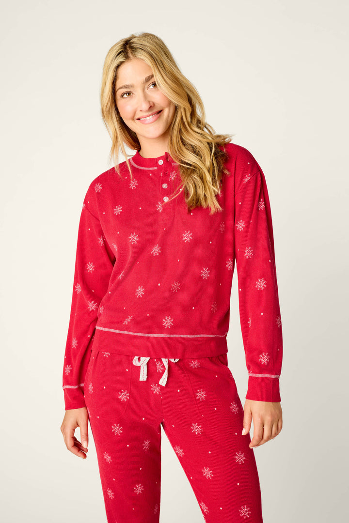 A person with long blonde hair smiles while wearing the P.J. Salvage Snowflakes Print 3-Piece PJ Gift Set, featuring a red pajama set with white snowflake patterns, against a plain light background.