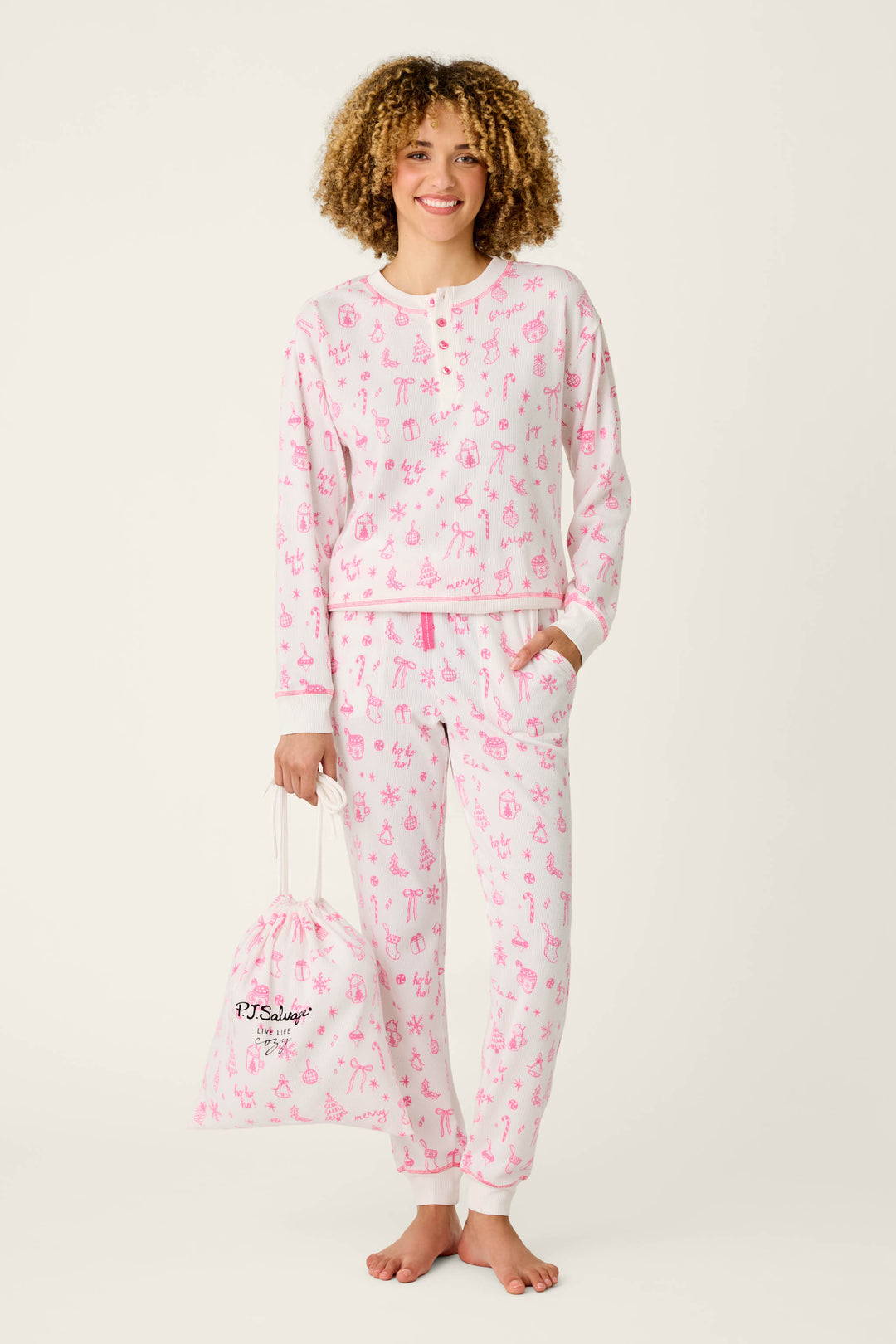 A smiling person with curly hair wears the P.J. Salvage Holiday Print 3-Piece PJ Gift Set, featuring matching white and pink pajamas adorned with playful pink animal and tree prints, and holds a coordinating white pajama bag. The background is plain and light-colored.