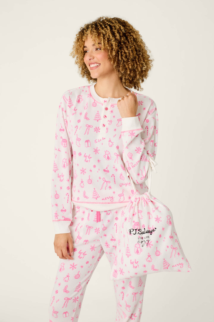 A smiling person with curly hair wears the P.J. Salvage Holiday Print 3-Piece PJ Gift Set—white pajamas featuring pink holiday prints—and holds a matching drawstring bag with “P.J. Salvage” in black, standing against a plain light background.