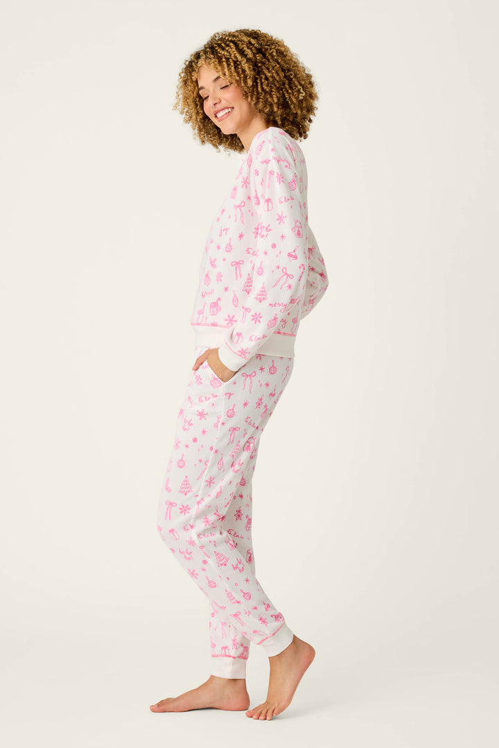 A person with curly hair smiles while standing barefoot in the cozy P.J. Salvage Holiday Print 3-Piece PJ Gift Set, featuring white pajamas adorned with pink holiday patterns like snowflakes and candy canes, set against a plain light background.