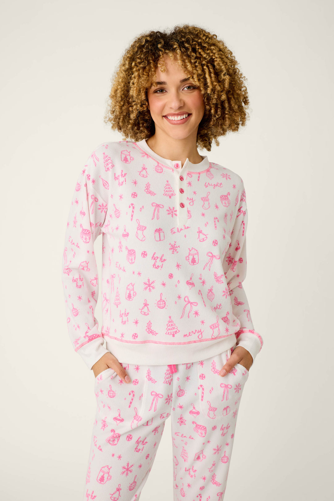 A person with curly hair smiles, wearing the P.J. Salvage Holiday Print 3-Piece PJ Gift Set—a matching white pajama set featuring pink holiday-themed drawings of reindeer, gifts, and trees—while standing against a plain light background.