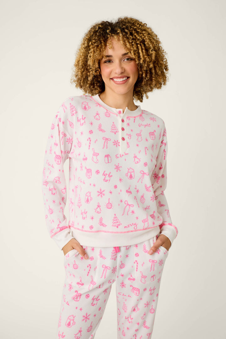 A person with curly hair smiles, wearing the P.J. Salvage Holiday Print 3-Piece PJ Gift Set—a matching white pajama set featuring pink holiday-themed drawings of reindeer, gifts, and trees—while standing against a plain light background.