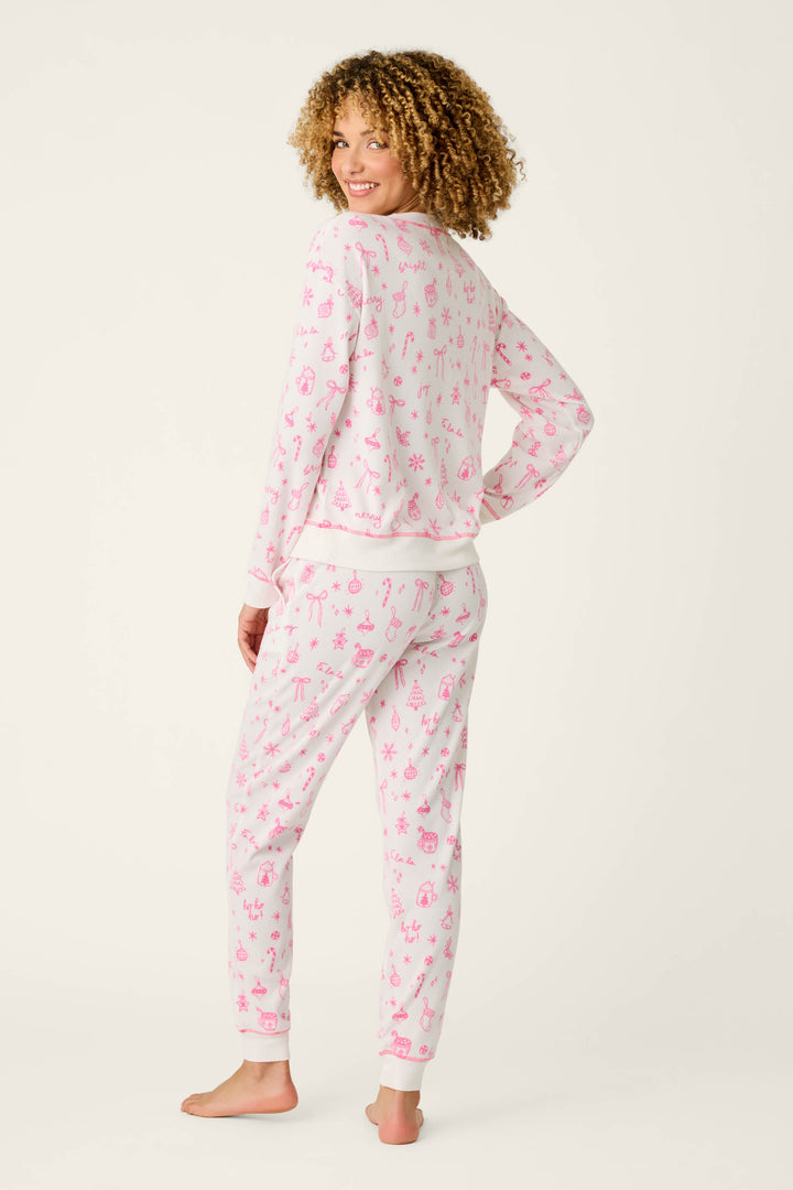A woman with curly hair stands barefoot and smiles over her shoulder, wearing the P.J. Salvage Holiday Print 3-Piece PJ Gift Set in a whimsical light pink animal print, set against a plain off-white background.