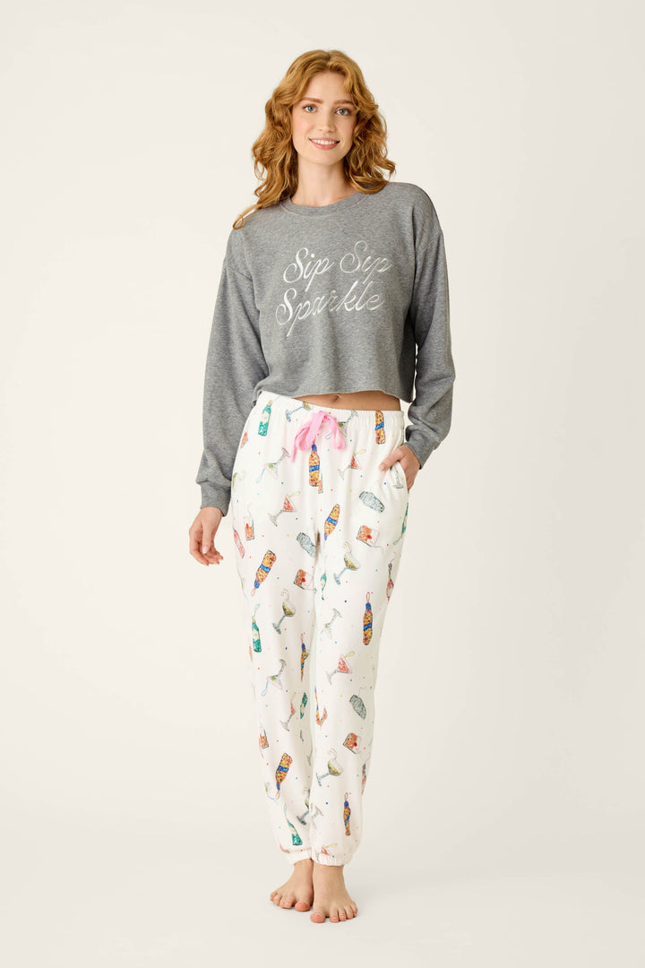 A woman wears a P.J. Salvage Sip Sip Sparkle Sweatshirt and white jogger pants featuring a colorful cocktail print.