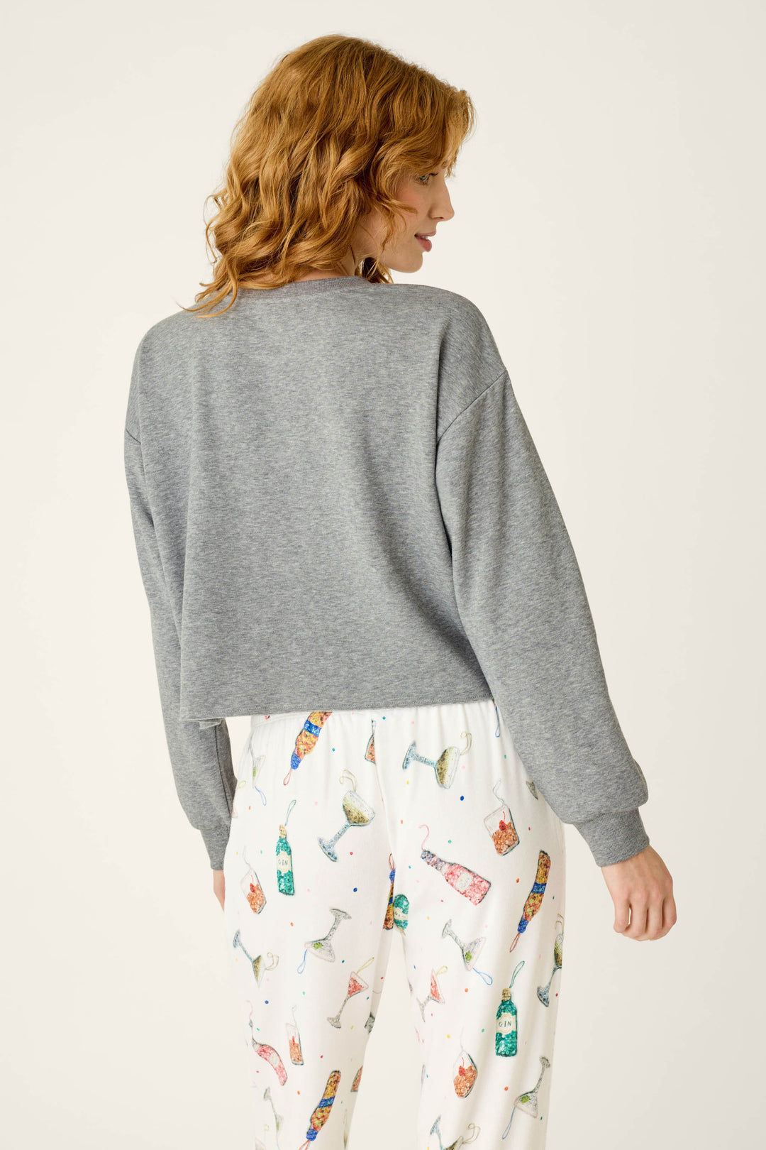 A person seen from behind wears a gray P.J. Salvage Sip Sip Sparkle Sweatshirt and white pajama pants patterned with colorful illustrations of cocktail glasses and bottles.