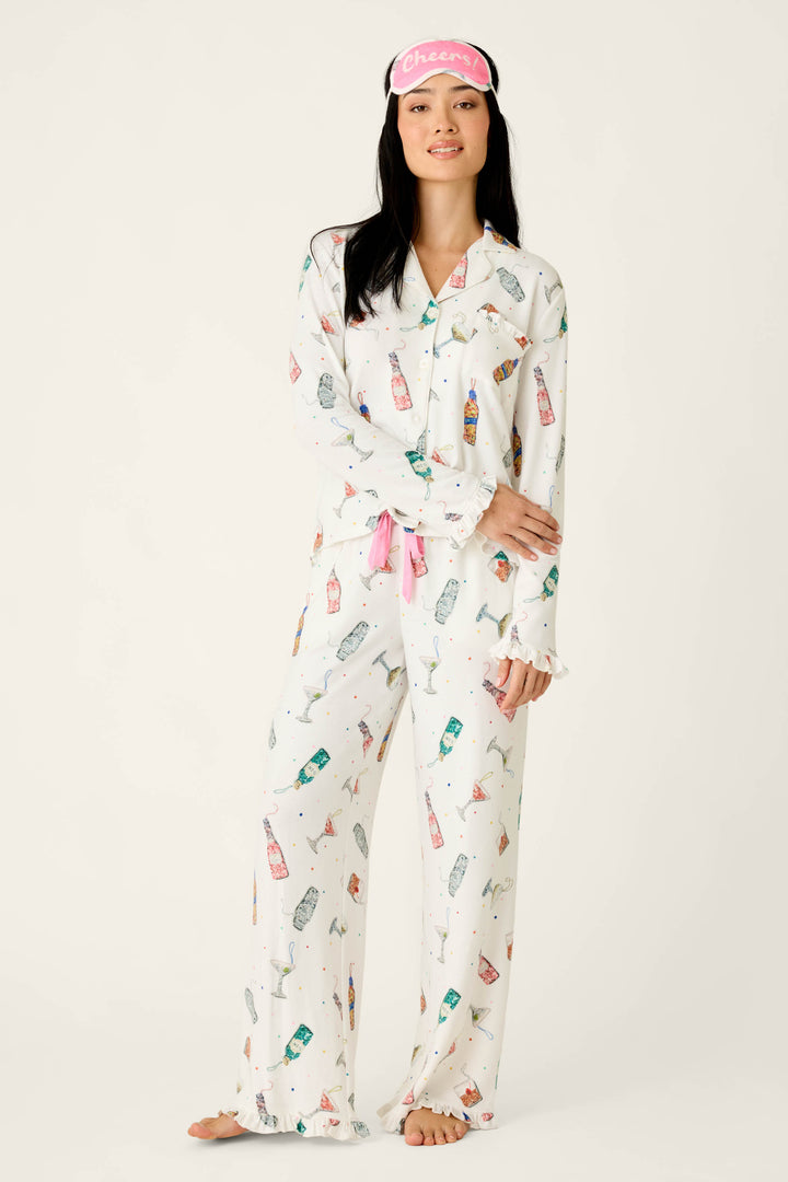 A woman wearing a Sip Sip Sparkle Cocktail Charms Print 3-Piece PJ Set in ivory with a confetti print and a pink sleep mask that reads Cheers.