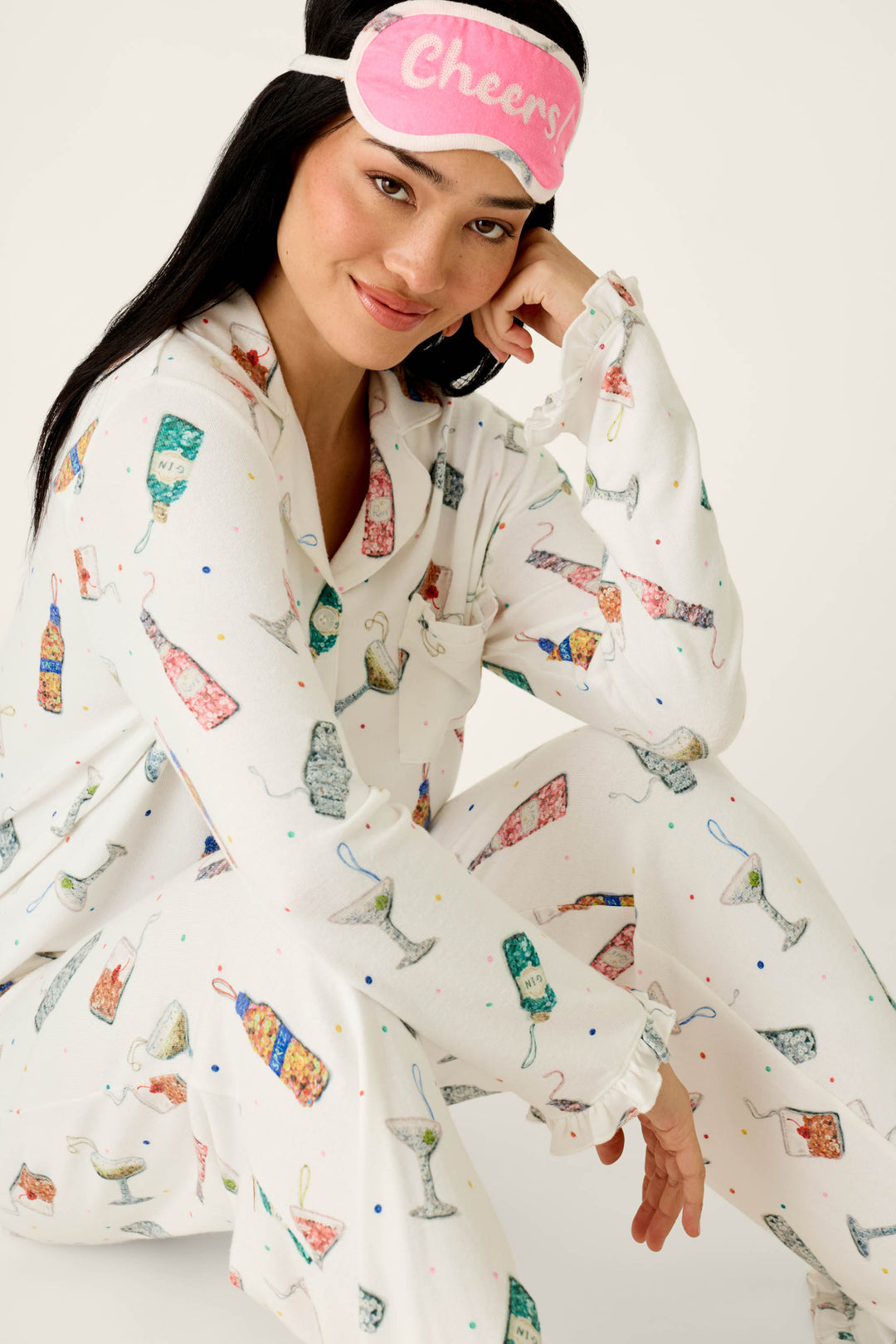 A woman wears a P.J. Salvage Sip Sip Sparkle Cocktail Charms Print 3-Piece PJ Set in ivory with a pink sleep mask that says Cheers!.