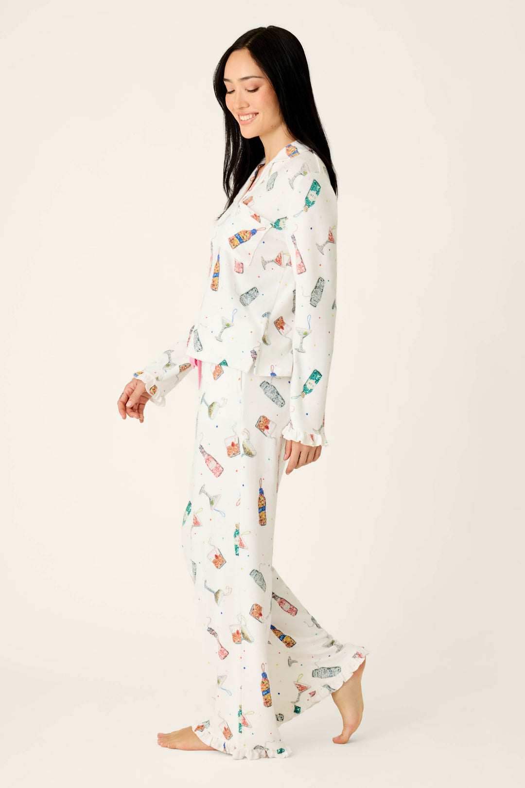 A woman wears the P.J. Salvage Sip Sip Sparkle Cocktail Charms Print 3-Piece PJ Set, an ivory pajama pant set featuring a colorful cocktail print and ruffled hems.