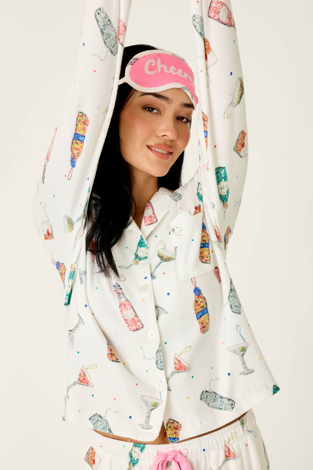 A smiling woman wears a pink sleep mask that says Cheers and the P.J. Salvage Sip Sip Sparkle Cocktail Charms Print 3-Piece PJ Set, featuring an ivory button-down top with a festive cocktail print.