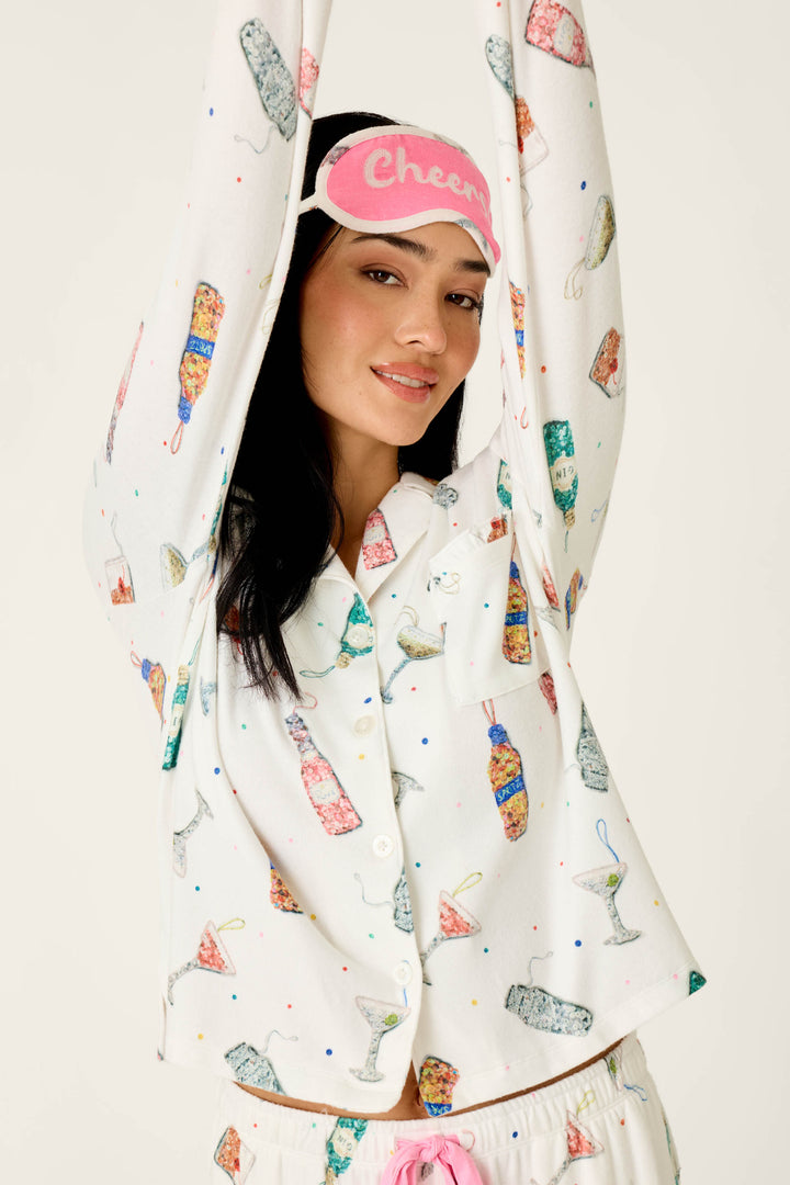 A smiling woman wears a pink sleep mask that says Cheers and the P.J. Salvage Sip Sip Sparkle Cocktail Charms Print 3-Piece PJ Set, featuring an ivory button-down top with a festive cocktail print.