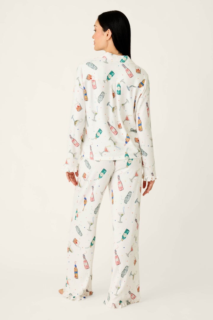 A person viewed from behind wearing a white Sip Sip Sparkle Cocktail Charms Print 3-Piece PJ Set featuring a colorful champagne bottle pattern and ruffled edges.