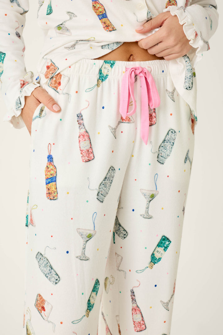 A person wearing the P.J. Salvage Sip Sip Sparkle Cocktail Charms Print 3-Piece PJ Set, featuring white pajamas with colorful cocktail bottles, martini glasses, scattered confetti dots, and a pink drawstring waist.