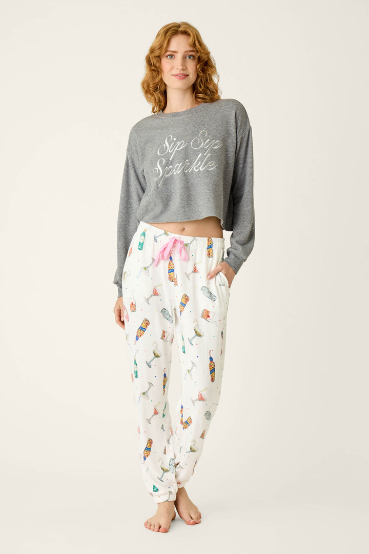 A woman wears a gray cropped sweatshirt with the text Sip Sip Sparkle and P.J. Salvage Sip Sip Sparkle Cocktail Charms Print Jogger Pant featuring a colorful cocktail pattern and a pink drawstring waist.