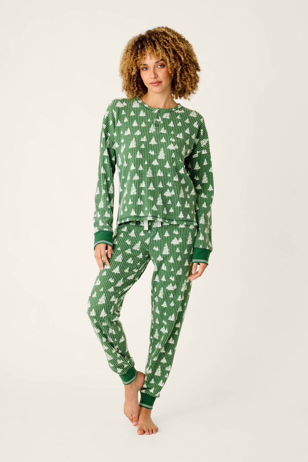 A person wearing a festive green and white Ski Jammie Classics Tree Print Long Sleeve PJ Set, featuring a thermal top and bottoms patterned with small trees.