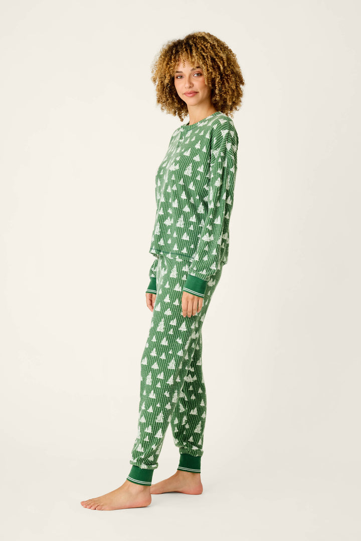 A woman wearing a festive green P.J. Salvage Ski Jammie Classics Tree Print Long Sleeve PJ Set featuring a white pine tree print.