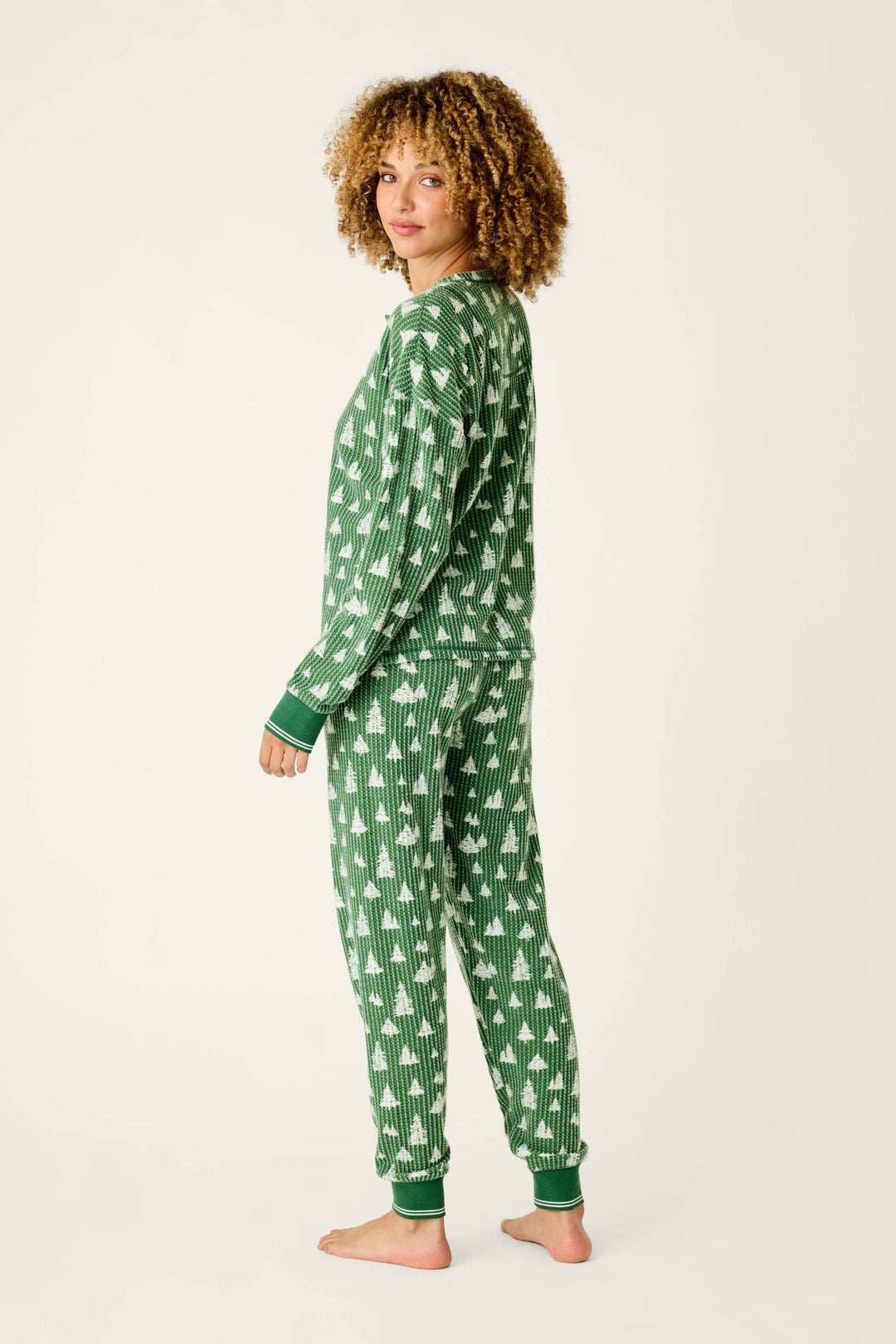 A person wearing a P.J. Salvage Ski Jammie Classics Tree Print Long Sleeve PJ Set, featuring a green waffle-knit long-sleeved top and matching jogger pants with a white pine tree pattern.
