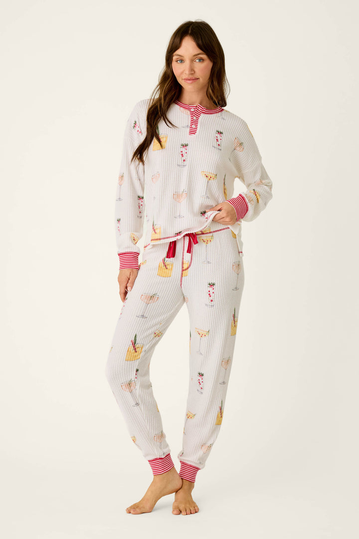 A woman wears a cozy P.J. Salvage Ski Jammie Classics Cocktail Print Long Sleeve PJ Set, featuring a cocktail print and red and white striped trim on the cuffs, neckline, and drawstring waist.