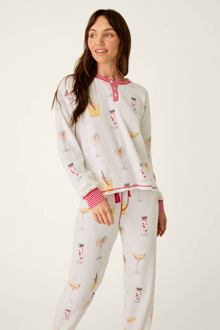 A woman wearing P.J. Salvage Ski Jammie Classics Cocktail Print Long Sleeve PJ Set, featuring a colorful cocktail pattern with red striped trim on the cuffs, neckline, and waistband.