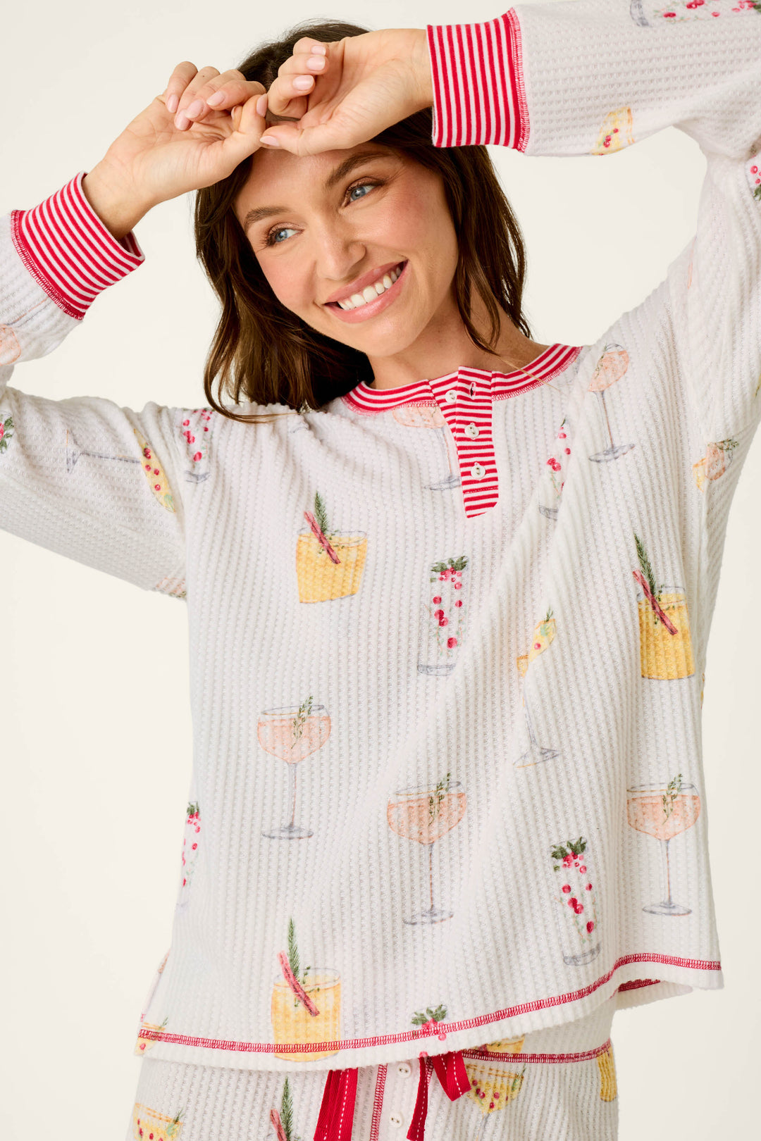 A woman wearing the P.J. Salvage Ski Jammie Classics Cocktail Print Long Sleeve PJ Set, a cozy thermal top featuring cocktail illustrations and red-and-white striped trim, while smiling.