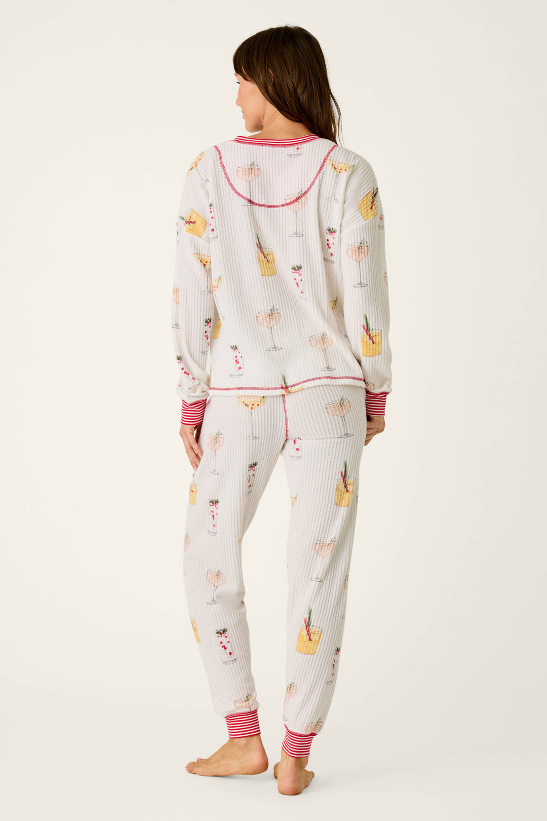 A woman viewed from the back wearing a P.J. Salvage Ski Jammie Classics Cocktail Print Long Sleeve PJ Set, featuring a white long-sleeve top and pants with a colorful cocktail print and red striped trim.