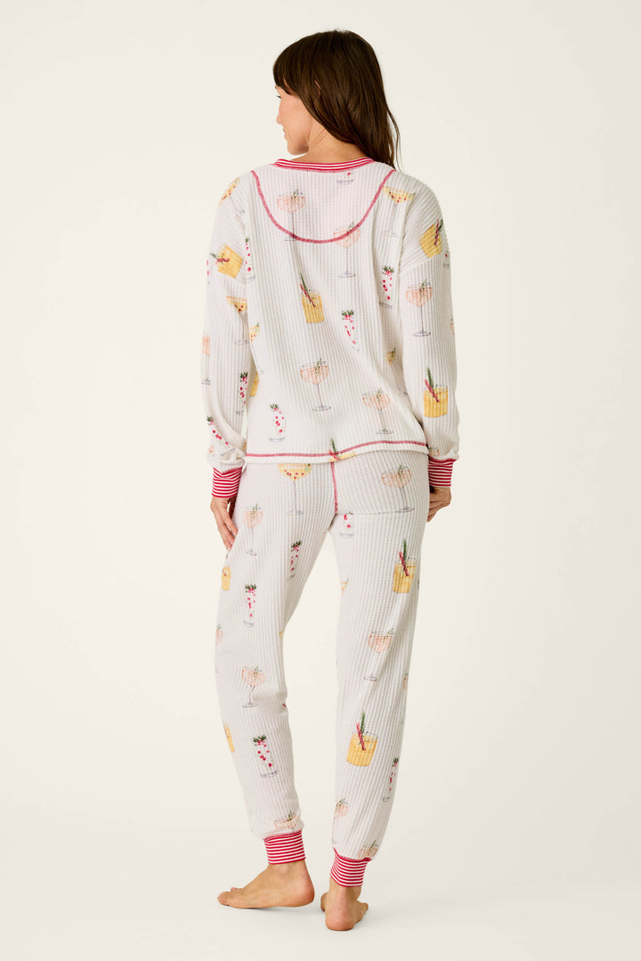 A woman viewed from the back wearing a P.J. Salvage Ski Jammie Classics Cocktail Print Long Sleeve PJ Set, featuring a white long-sleeve top and pants with a colorful cocktail print and red striped trim.