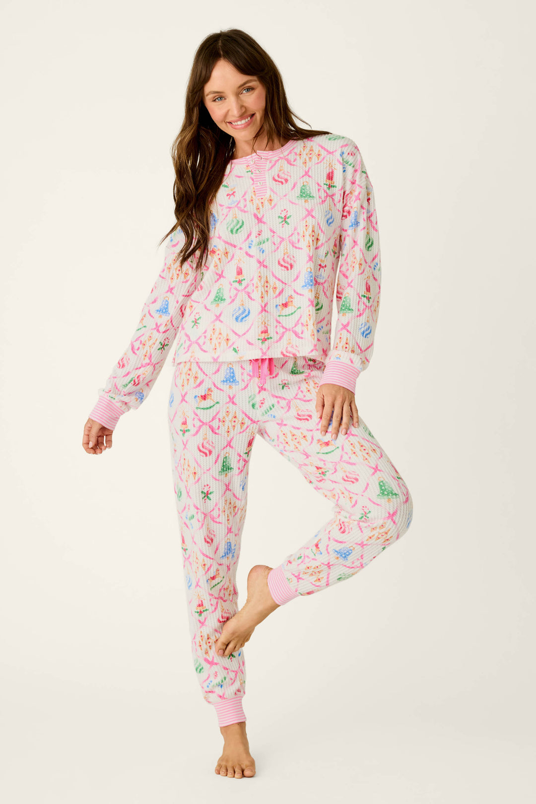 A smiling woman wears P.J. Salvage Ski Jammie Classics Ornament Print Long Sleeve PJ Set, a festive waffle-knit pajama set with a white background and a colorful print of Christmas ornaments and ribbons.