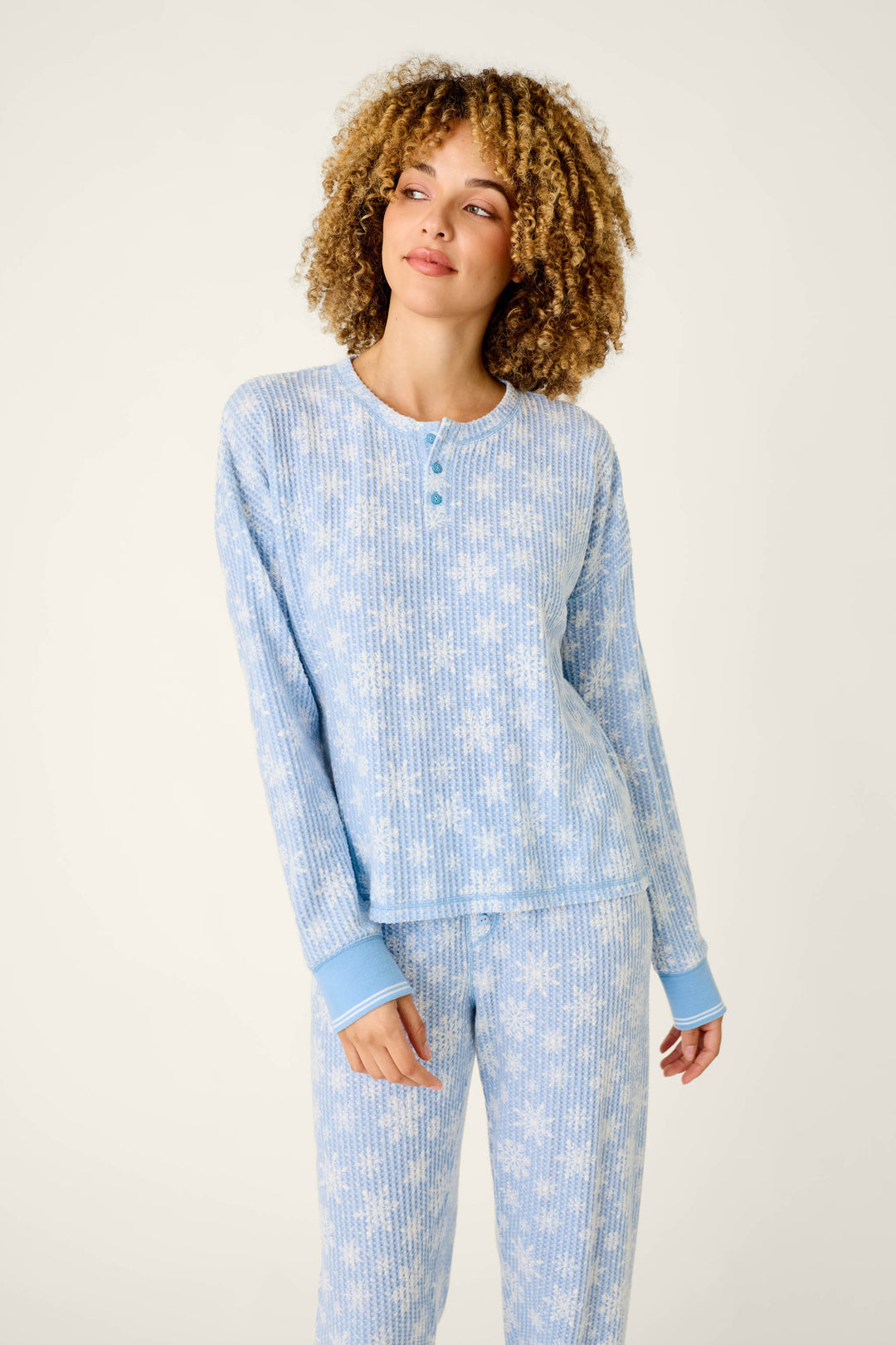 A woman wears a light blue P.J. Salvage Ski Jammie Classics Snowflake Print Long Sleeve PJ Set, featuring a waffle-knit texture and a white snowflake pattern on the long-sleeve henley top and matching pants.