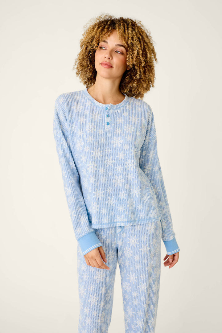 A woman wears a light blue P.J. Salvage Ski Jammie Classics Snowflake Print Long Sleeve PJ Set, featuring a waffle-knit texture and a white snowflake pattern on the long-sleeve henley top and matching pants.