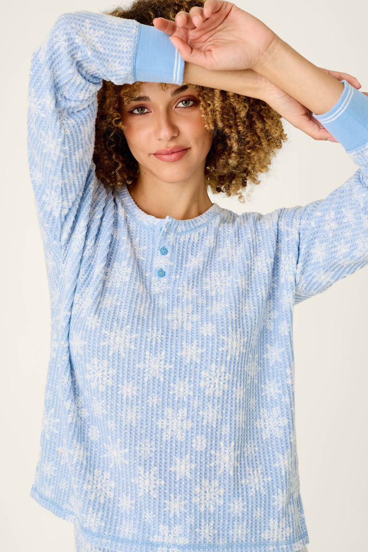 A woman with curly hair wears a light blue brushed thermal P.J. Salvage Ski Jammie Classics Snowflake Print Long Sleeve PJ Set henley shirt featuring a white snowflake pattern and solid blue cuffs.