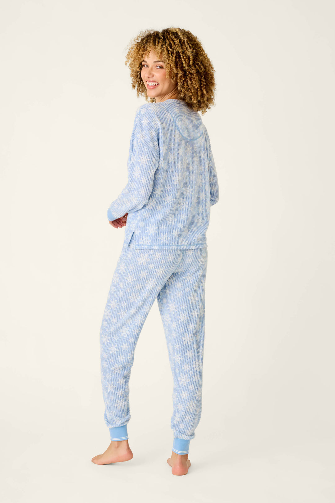 A woman wearing a P.J. Salvage Ski Jammie Classics Snowflake Print Long Sleeve PJ Set, featuring a light blue top and pants with a white snowflake pattern and ribbed cuffs.