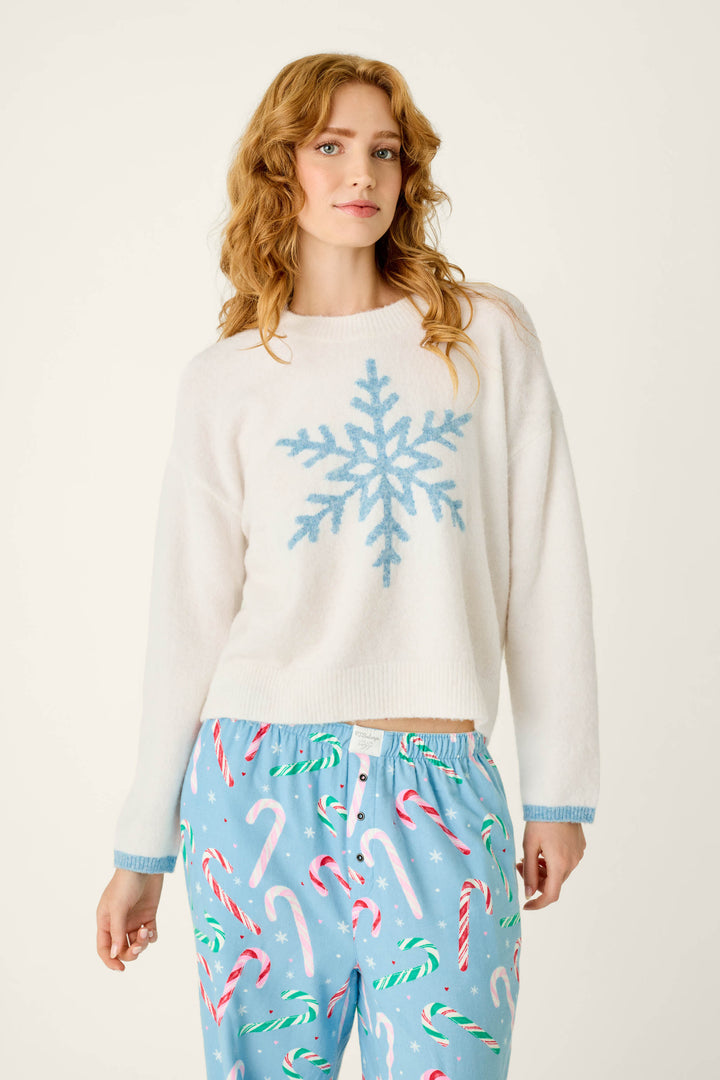 A model wears a P.J. Salvage The Sweetest Season Snowflake Snuggle Sweater, a cozy white long-sleeve top with a blue snowflake graphic paired with light blue pajama pants patterned with candy canes and small snowflakes.