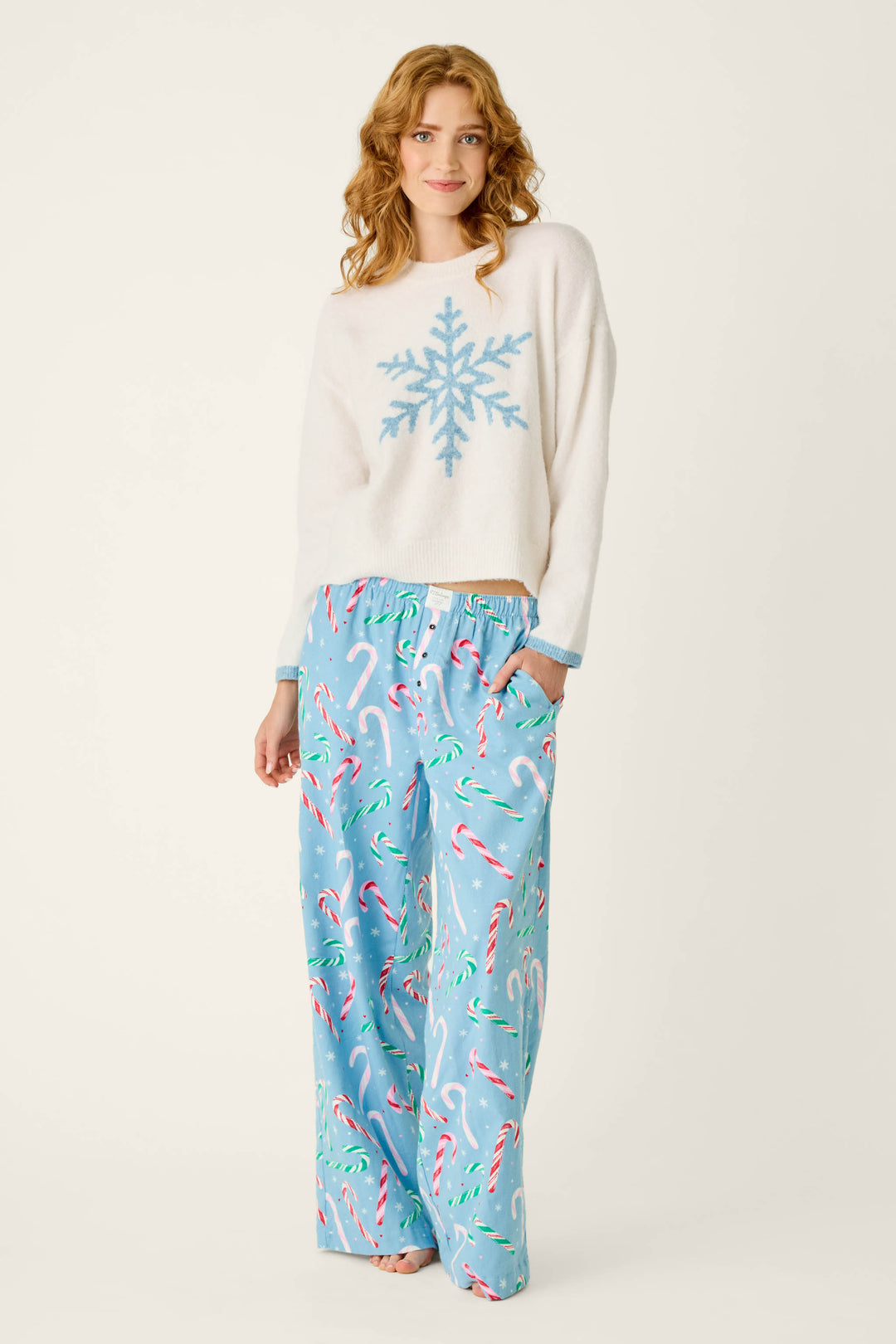 A woman wearing a P.J. Salvage The Sweetest Season Snowflake Snuggle Sweater, a cream-colored top with a large blue snowflake design, paired with matching blue pants featuring a candy cane and snowflake print.