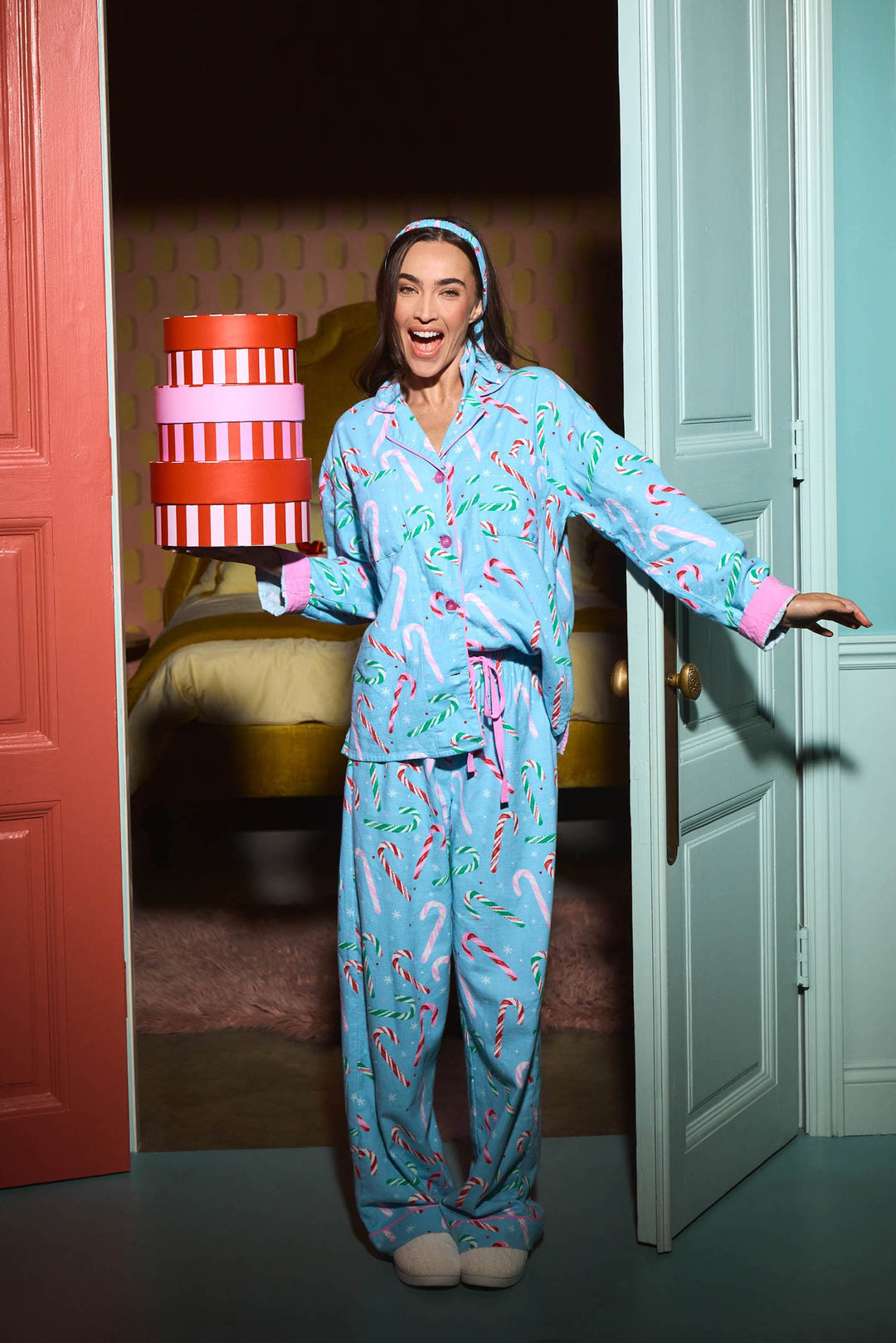 A woman wears a light blue The Sweetest Season Candy Cane Print Flannel 3-Piece PJ Set by P.J. Salvage as she holds a stack of three gift boxes in a doorway.