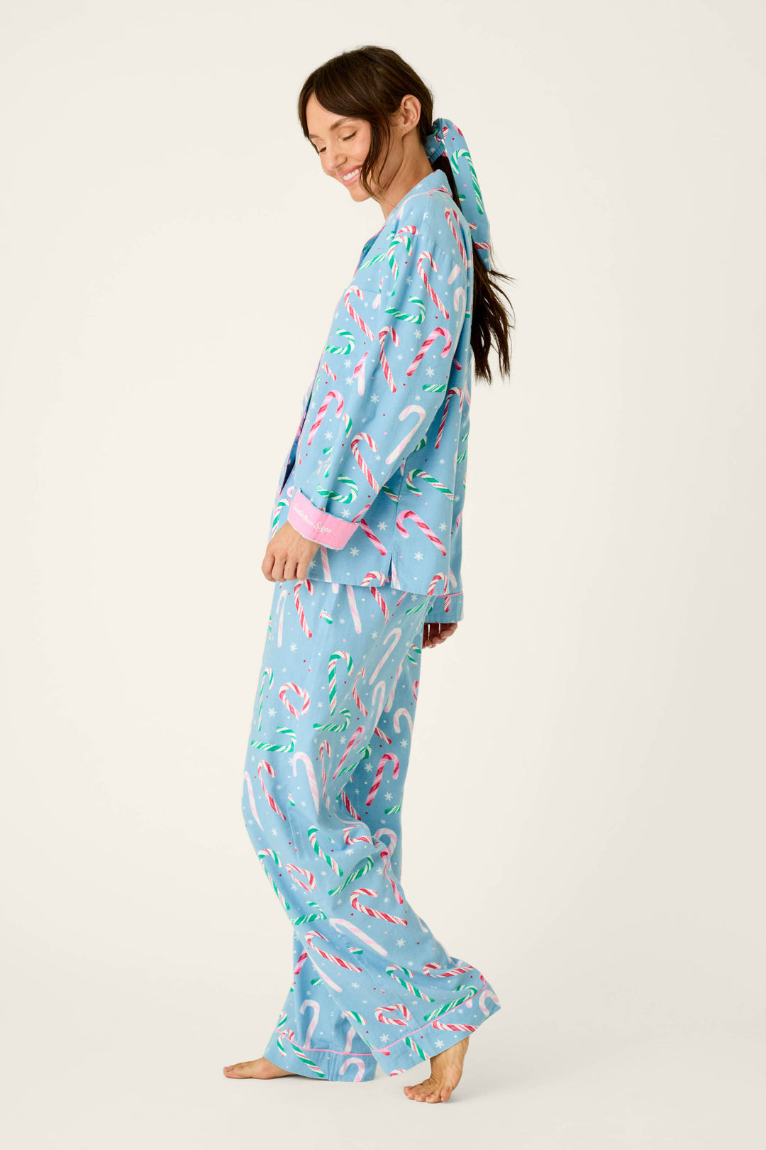 A person wearing a light blue The Sweetest Season Candy Cane Print Flannel 3-Piece PJ Set featuring a candy cane and snowflake print, including a button-down top, pants, and a matching hair scrunchie.