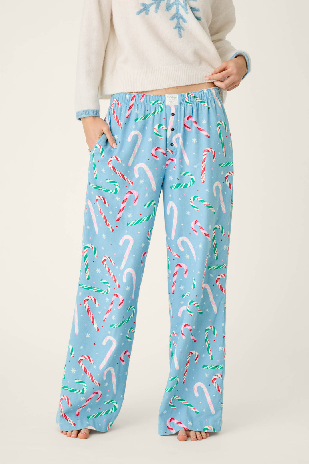 A person wears P.J. Salvage The Sweetest Season Candy Cane Print Flannel Pajama Pants in cozy, light blue fabric featuring a festive print of red, white, and green candy canes and small white snowflakes.