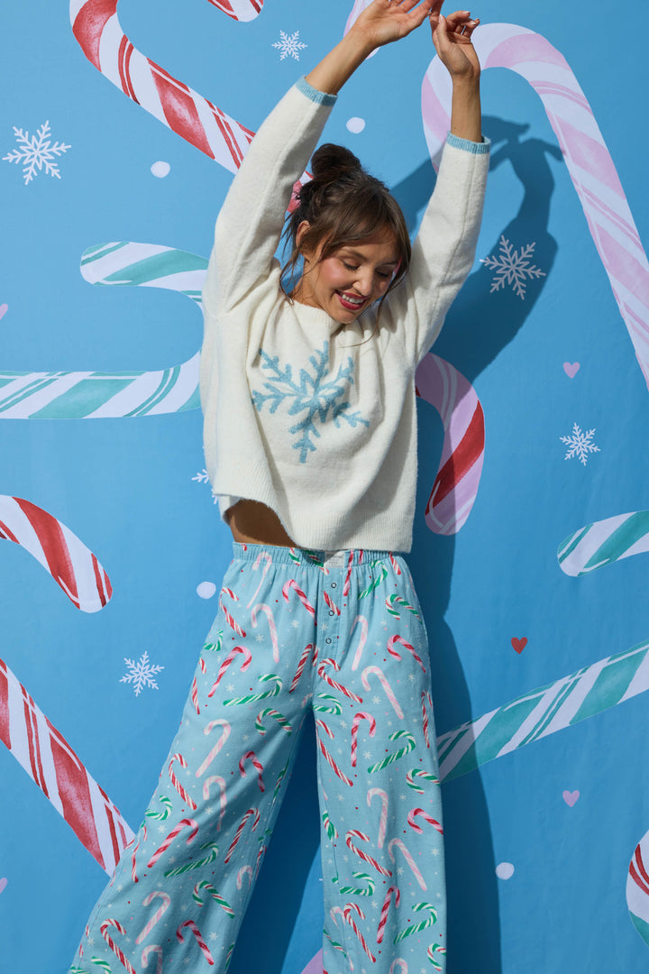 A woman in a cream-colored snowflake sweater and light blue The Sweetest Season Candy Cane Print Flannel Pajama Pants poses with her arms raised against a blue background decorated with large candy canes and snowflakes.