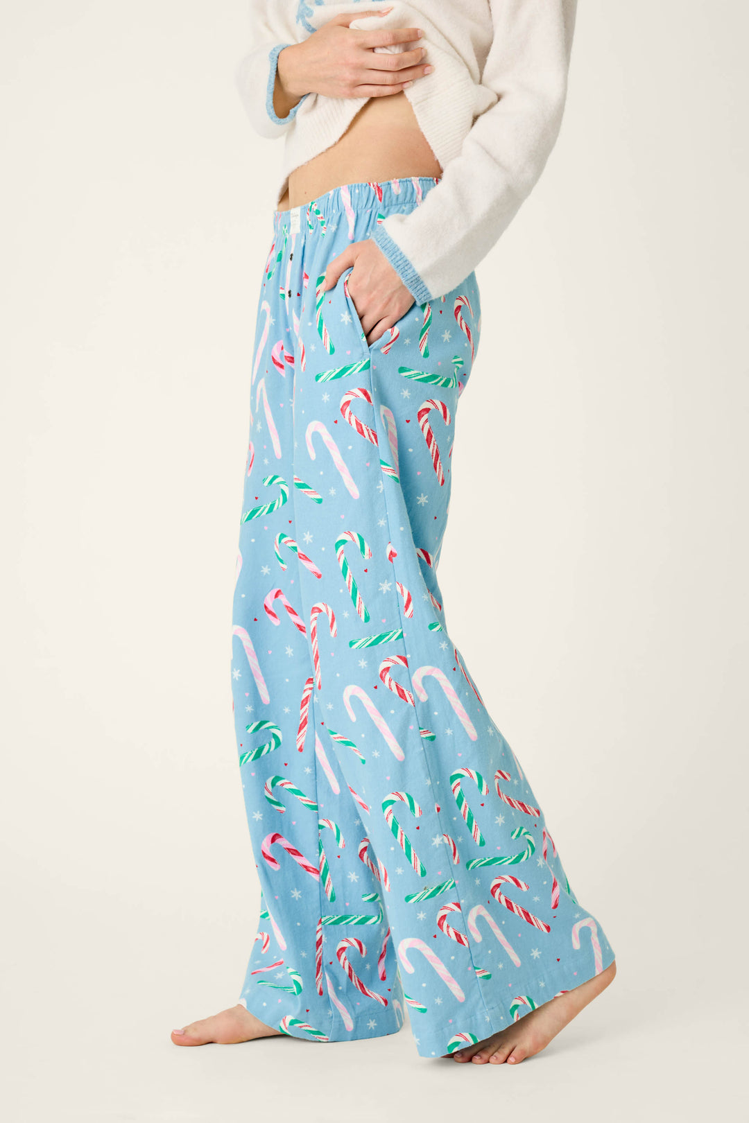 A person in a white sweater wearing cozy, light blue The Sweetest Season Candy Cane Print Flannel Pajama Pants featuring a festive candy cane and snowflake print.