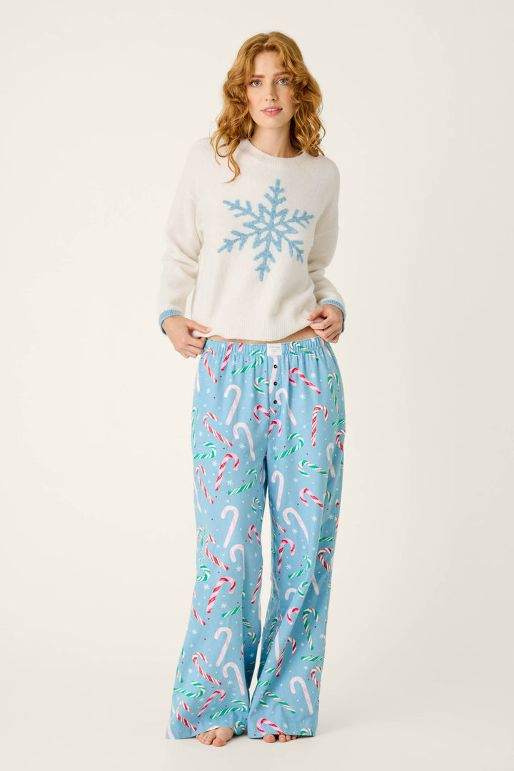A woman wears a cream-colored sweater with a blue snowflake design and P.J. Salvage The Sweetest Season Candy Cane Print Flannel Pajama Pants featuring a candy cane and white snowflake pattern.
