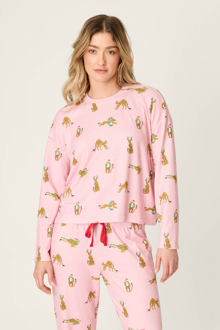 A woman wearing a pink P.J. Salvage Very Merry Wild for the Holidays Long Sleeve Top and matching bottoms patterned with small leopards wearing holiday hats and accessories.