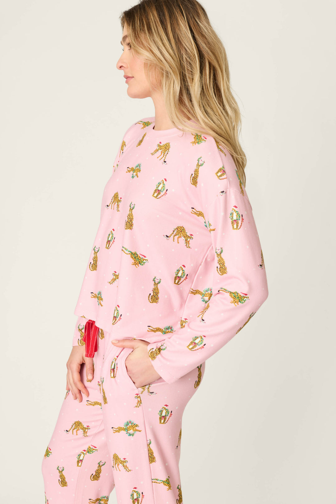 A woman wears a P.J. Salvage Very Merry Wild for the Holidays Long Sleeve Top and matching pants, both light pink and covered in a pattern of leopards wearing Christmas hats and scarves.