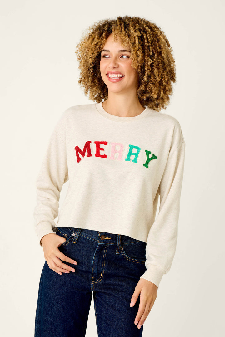 A smiling person wears a cozy, cream-colored P.J. Salvage Very Merry Bouclé Merry Sweatshirt with MERRY in colorful, textured embroidery on the chest.