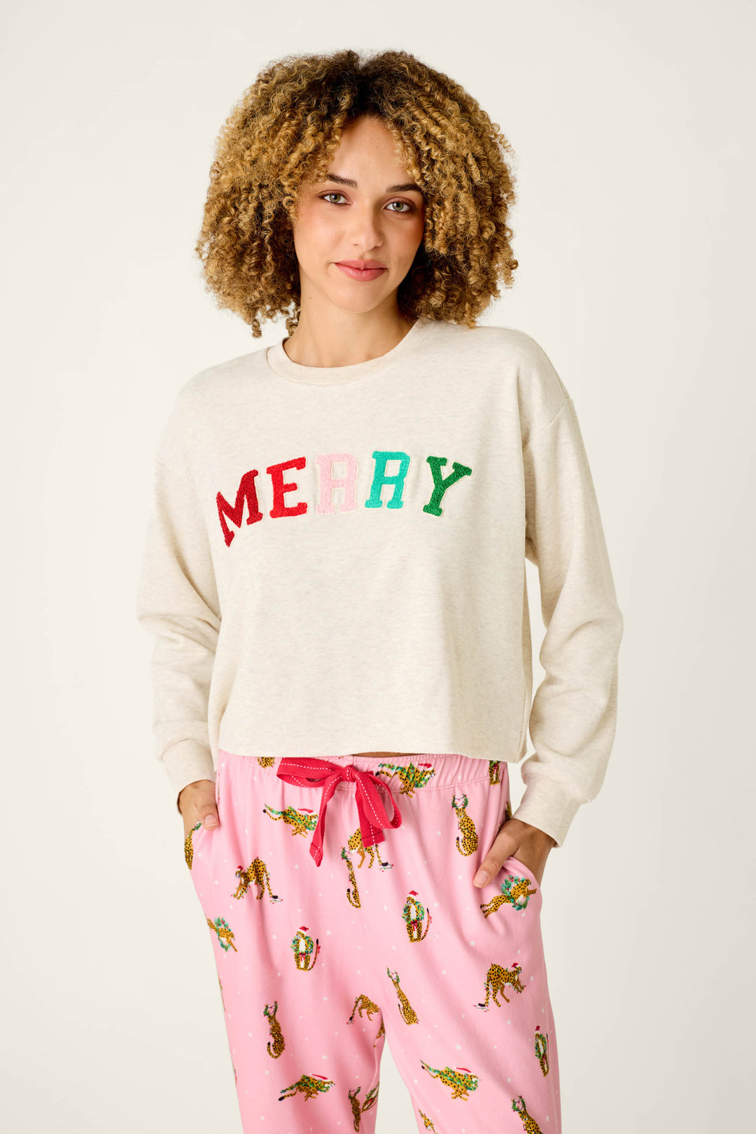 A person wearing a P.J. Salvage Very Merry Bouclé Merry Sweatshirt in cream with colorful, textured letters, paired with pink leopard-print pajama pants.